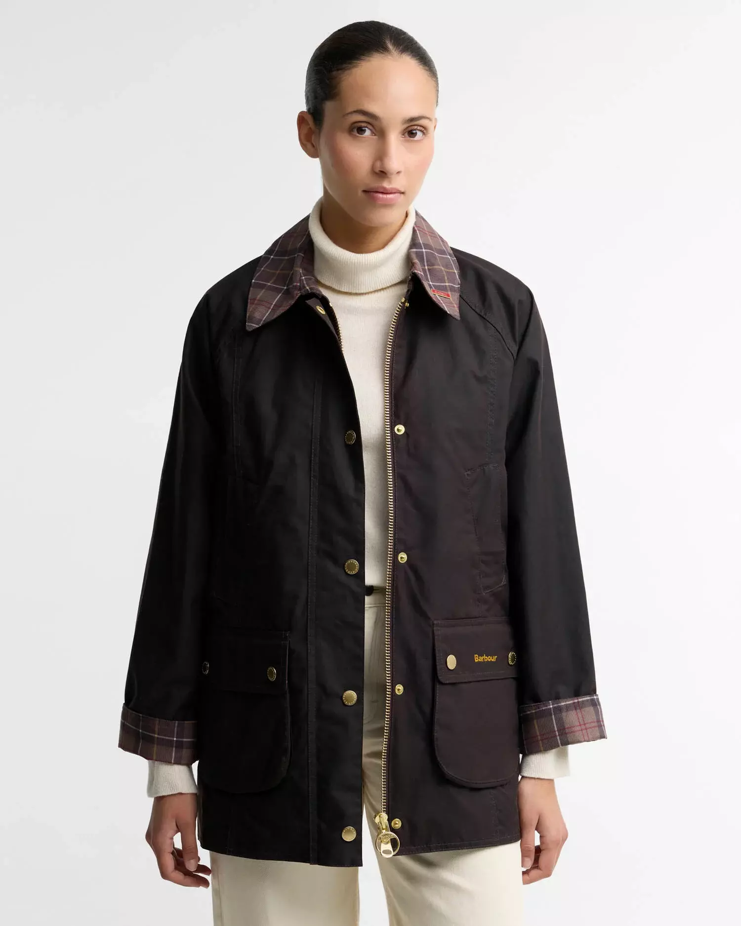 Woman in a dark Barbour wax jacket with plaid collar, gold zipper, and cream turtleneck, facing forward on white background