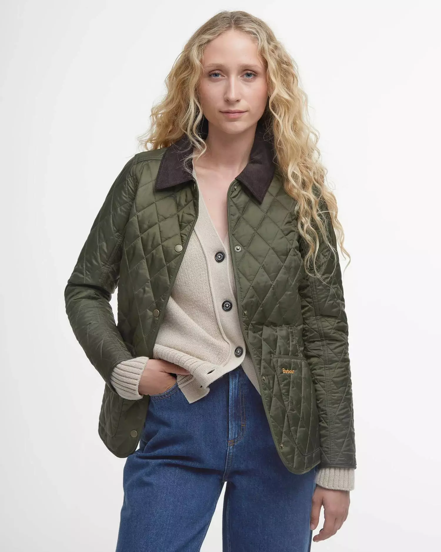 Young woman models a green quilted Barbour jacket with a brown corduroy collar, beige cardigan, blue jeans